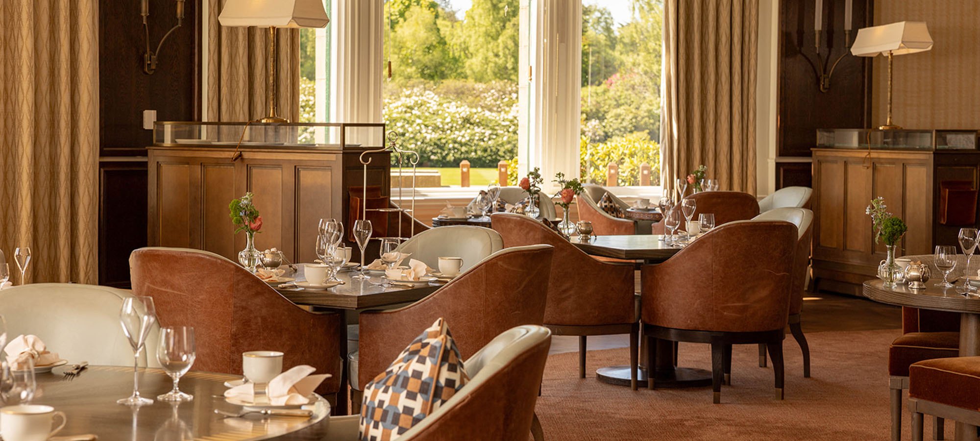 Leather tub chairs and tables laid for tea in a Gleneagles lounge, the gardens through the window